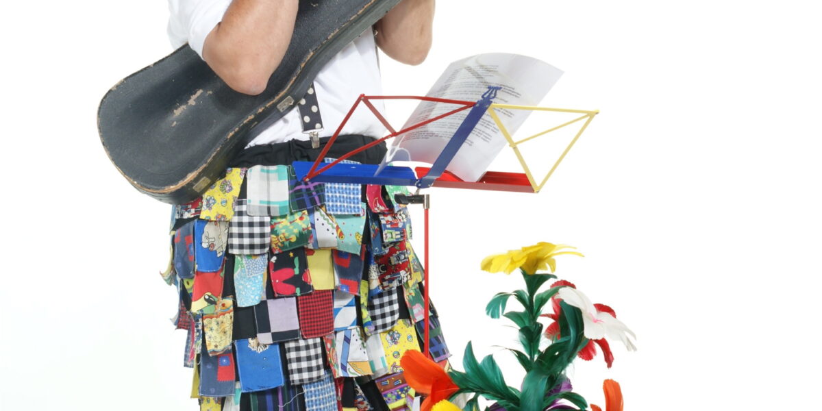Ein Clown in bunten Patchworkhosen und einem hohen Hut spielt Flöte und hält einen Geigenkasten in der Hand. Er steht neben einer Topfpflanze mit künstlichen Blumen und einem Notenständer.