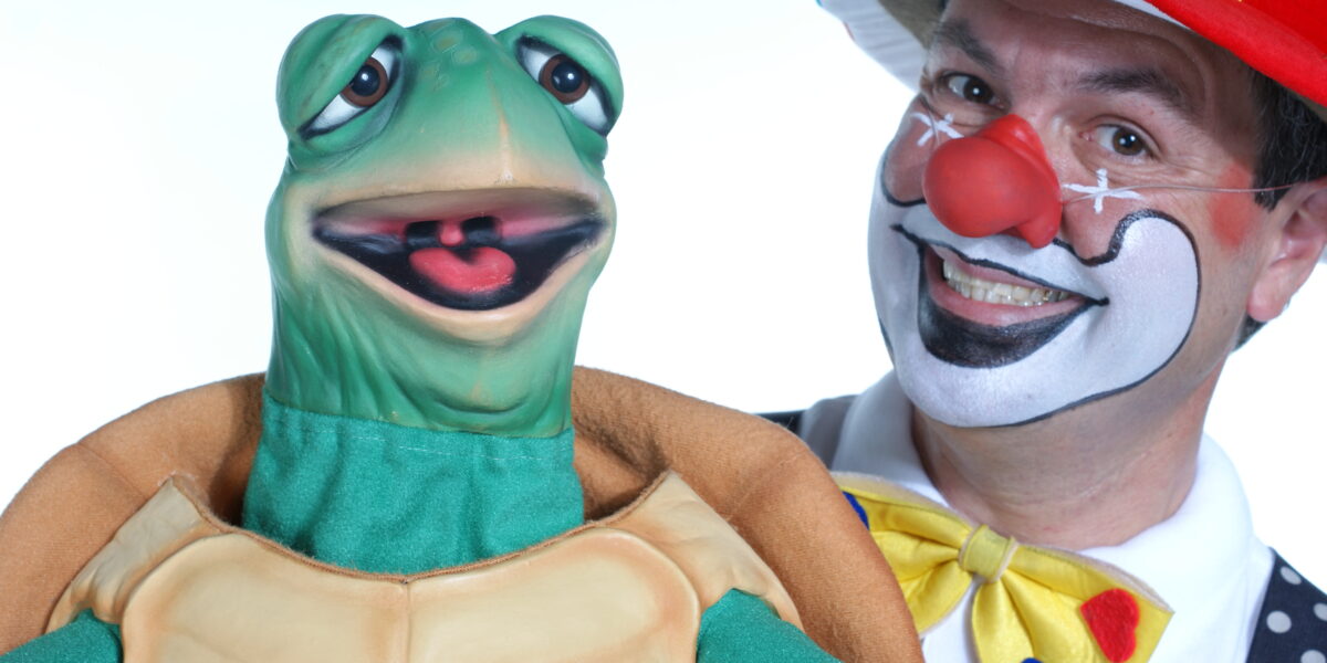 Eine als Clown verkleidete Person mit Gesichtsbemalung und einem bunten Hut steht neben einer grünen Schildkrötenpuppe, beide lächeln in die Kamera.