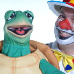 Eine als Clown verkleidete Person mit Gesichtsbemalung und einem bunten Hut steht neben einer grünen Schildkrötenpuppe, beide lächeln in die Kamera.