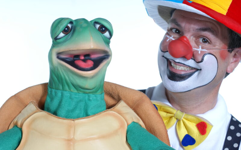 Eine als Clown verkleidete Person mit Gesichtsbemalung und einem bunten Hut steht neben einer grünen Schildkrötenpuppe, beide lächeln in die Kamera.