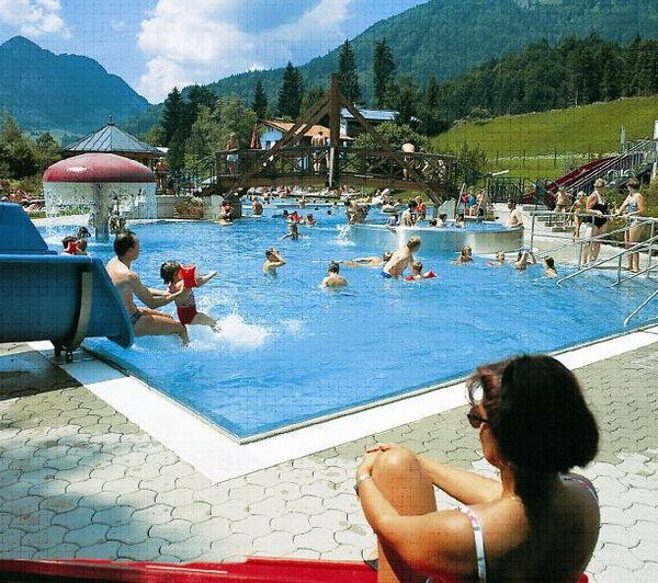 Menschen schwimmen und spielen in einem großen Freibad, umgeben von Bergen und Bäumen, während eine Frau am Rand sitzt und die Szene beobachtet.