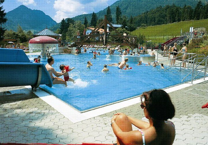 Menschen schwimmen und spielen in einem großen Freibad, umgeben von Bergen und Bäumen, während eine Frau am Rand sitzt und die Szene beobachtet.