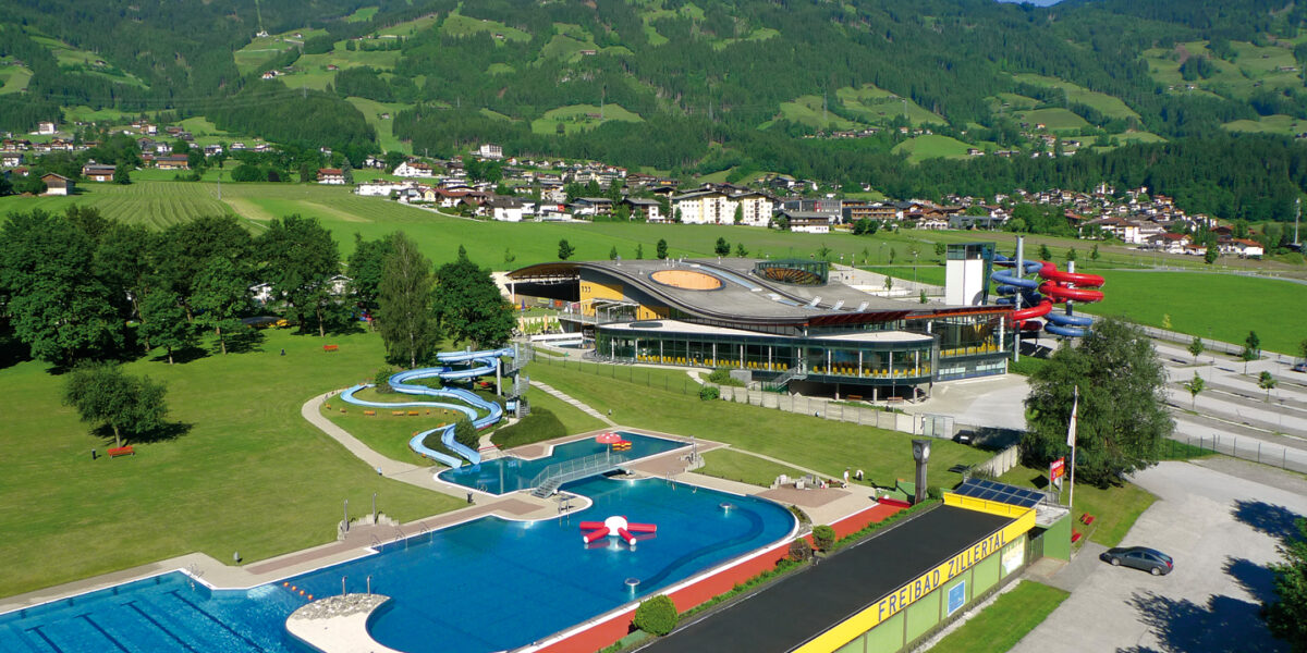 Ein großer Freibadkomplex mit Rutschen und einem modernen Gebäude, umgeben von grünen Feldern, Bäumen und einer Kulisse aus bewaldeten Hügeln und einer kleinen Stadt.