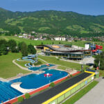 Ein großer Freibadkomplex mit Rutschen und einem modernen Gebäude, umgeben von grünen Feldern, Bäumen und einer Kulisse aus bewaldeten Hügeln und einer kleinen Stadt.