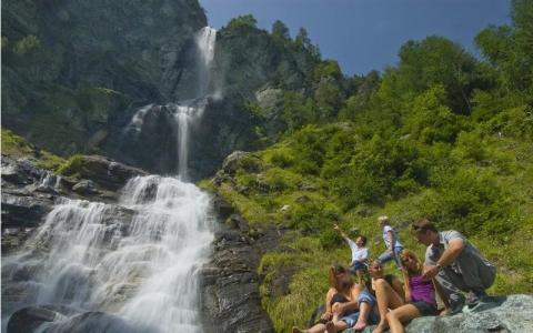 Eine Gruppe von Menschen sitzt auf grasbewachsenen Felsen in der Nähe des Fußes eines hohen Wasserfalls, umgeben von grünen Bäumen und steilen Felsen unter blauem Himmel.