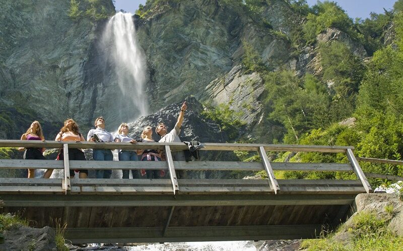 Eine Gruppe von Menschen steht auf einer Holzbrücke und macht ein Selfie, mit einem Wasserfall und grünen Bäumen im Hintergrund.