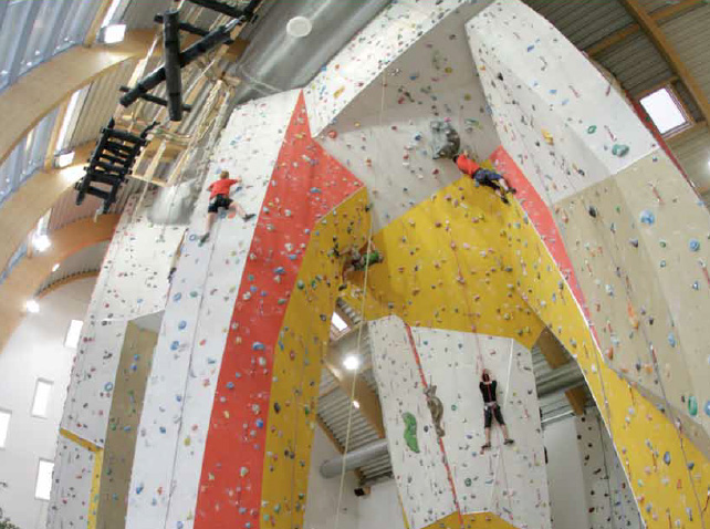 In einer großen, gut beleuchteten Anlage klettern mehrere Personen an hohen Indoor-Kletterwänden mit bunten Griffen.