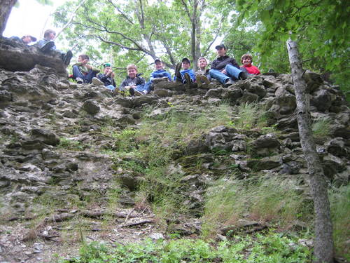 Eine Gruppe von Menschen sitzt am Rande einer felsigen Klippe und blickt nach unten, umgeben von Bäumen und Grünzeug.