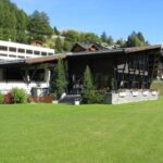 Modernes Gebäude mit kantigem Dach und Glaswänden auf einer Rasenfläche, umgeben von Bäumen und nahegelegenen Chaletbauten unter einem klaren blauen Himmel.