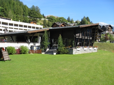 Modernes Gebäude mit kantigem Dach und Glaswänden auf einer Rasenfläche, umgeben von Bäumen und nahegelegenen Chaletbauten unter einem klaren blauen Himmel.