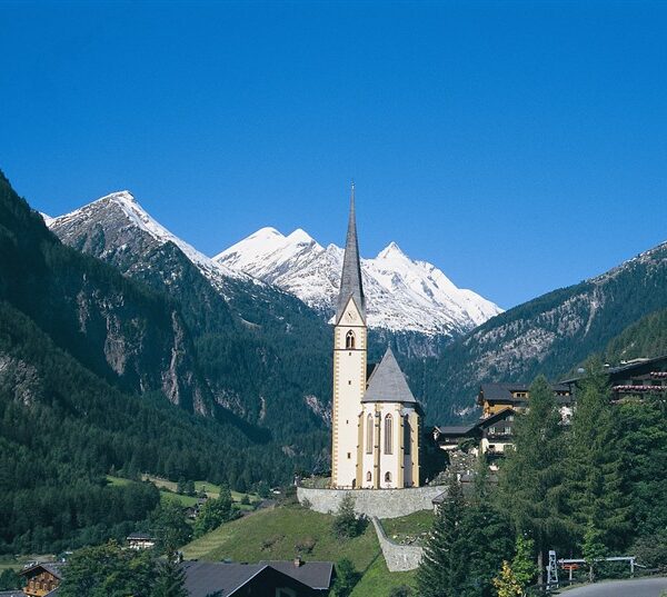 Eine hohe Kirche mit spitzem Kirchturm steht in einem grünen Tal, umgeben von Alpenhäusern und bewaldeten Bergen, mit schneebedeckten Gipfeln im Hintergrund.