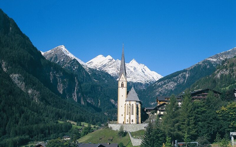 Eine hohe Kirche mit spitzem Kirchturm steht in einem grünen Tal, umgeben von Alpenhäusern und bewaldeten Bergen, mit schneebedeckten Gipfeln im Hintergrund.