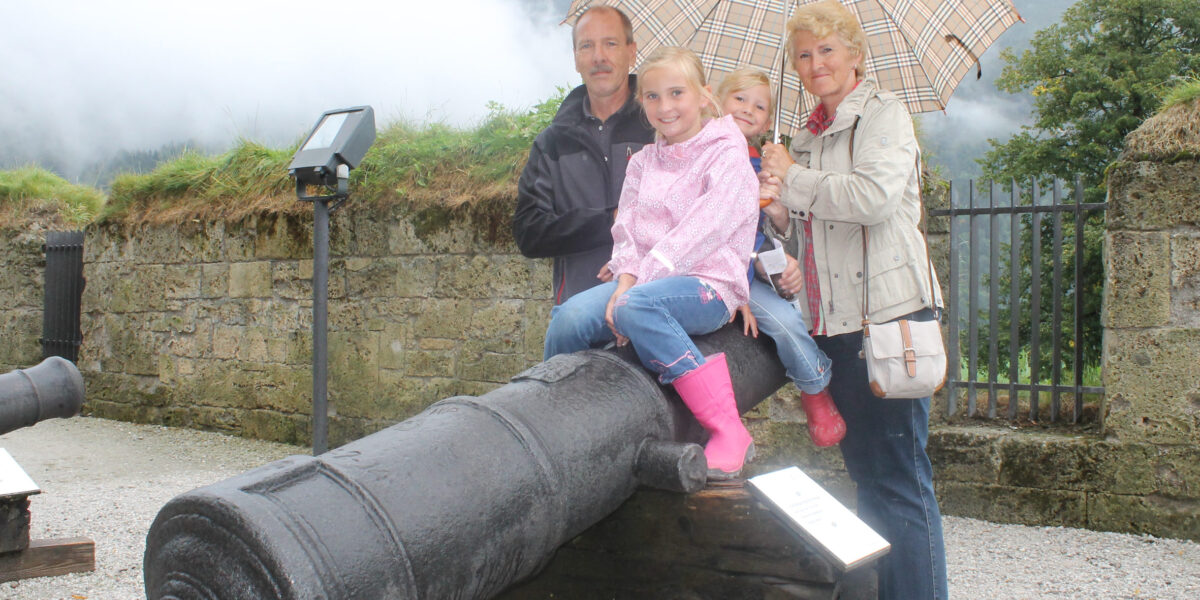 Eine vierköpfige Familie posiert an einem bewölkten Tag im Freien an einer alten Kanone, wobei zwei Kinder auf der Kanone sitzen und zwei Erwachsene hinter ihnen stehen und einen Regenschirm halten.