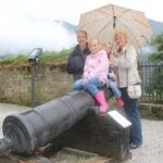 Eine vierköpfige Familie posiert an einem bewölkten Tag im Freien an einer alten Kanone, wobei zwei Kinder auf der Kanone sitzen und zwei Erwachsene hinter ihnen stehen und einen Regenschirm halten.