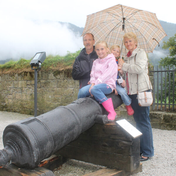 Eine vierköpfige Familie posiert an einem bewölkten Tag im Freien an einer alten Kanone, wobei zwei Kinder auf der Kanone sitzen und zwei Erwachsene hinter ihnen stehen und einen Regenschirm halten.
