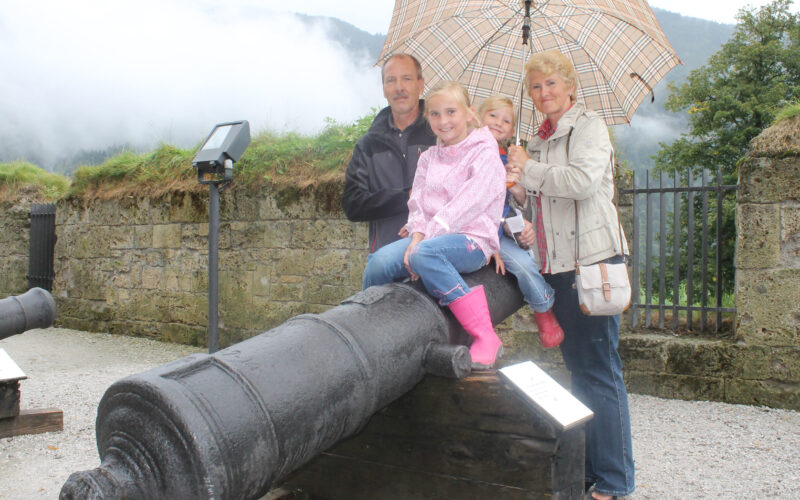 Eine vierköpfige Familie posiert an einem bewölkten Tag im Freien an einer alten Kanone, wobei zwei Kinder auf der Kanone sitzen und zwei Erwachsene hinter ihnen stehen und einen Regenschirm halten.