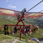 Vier Menschen in Gurten schweben an einem klaren, sonnigen Tag mit einem Seilrutschen-ähnlichen Gleitschirm über grasbewachsene Hügel, heben die Arme und blicken in die Kamera.