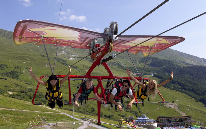 Vier Menschen in Gurten schweben an einem klaren, sonnigen Tag mit einem Seilrutschen-ähnlichen Gleitschirm über grasbewachsene Hügel, heben die Arme und blicken in die Kamera.