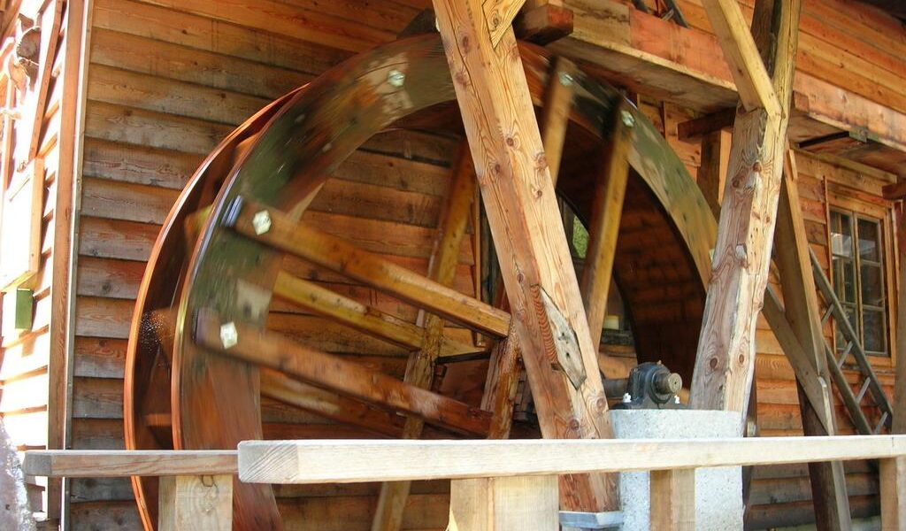 Ein großes hölzernes Wasserrad dreht sich neben einem Holzgebäude, das von Balken getragen wird und sich auf einer Achse dreht.