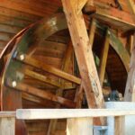 Ein großes hölzernes Wasserrad dreht sich neben einem Holzgebäude, das von Balken getragen wird und sich auf einer Achse dreht.