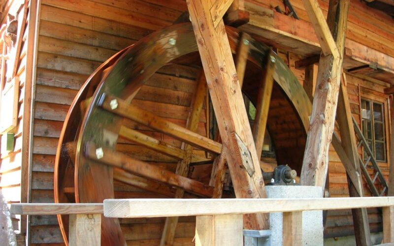 Ein großes hölzernes Wasserrad dreht sich neben einem Holzgebäude, das von Balken getragen wird und sich auf einer Achse dreht.