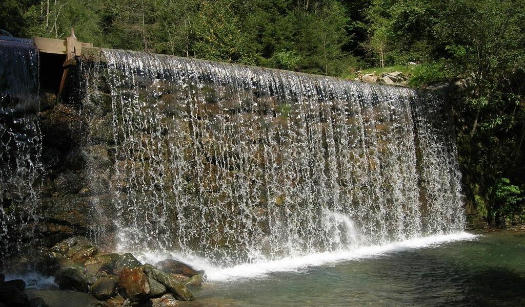 Ein künstlich angelegter Wasserfall fließt über einen von Bäumen umgebenen Stein- und Betondamm und stürzt in ein klares Wasserbecken.