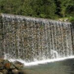 Ein künstlich angelegter Wasserfall fließt über einen von Bäumen umgebenen Stein- und Betondamm und stürzt in ein klares Wasserbecken.