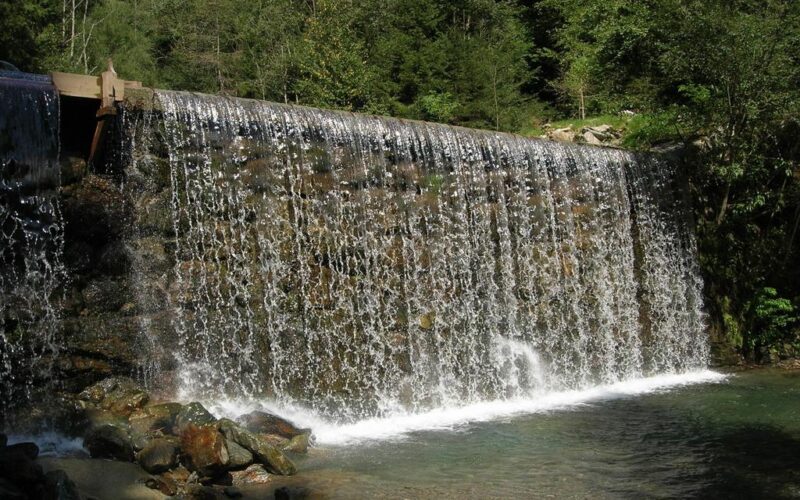 Ein künstlich angelegter Wasserfall fließt über einen von Bäumen umgebenen Stein- und Betondamm und stürzt in ein klares Wasserbecken.