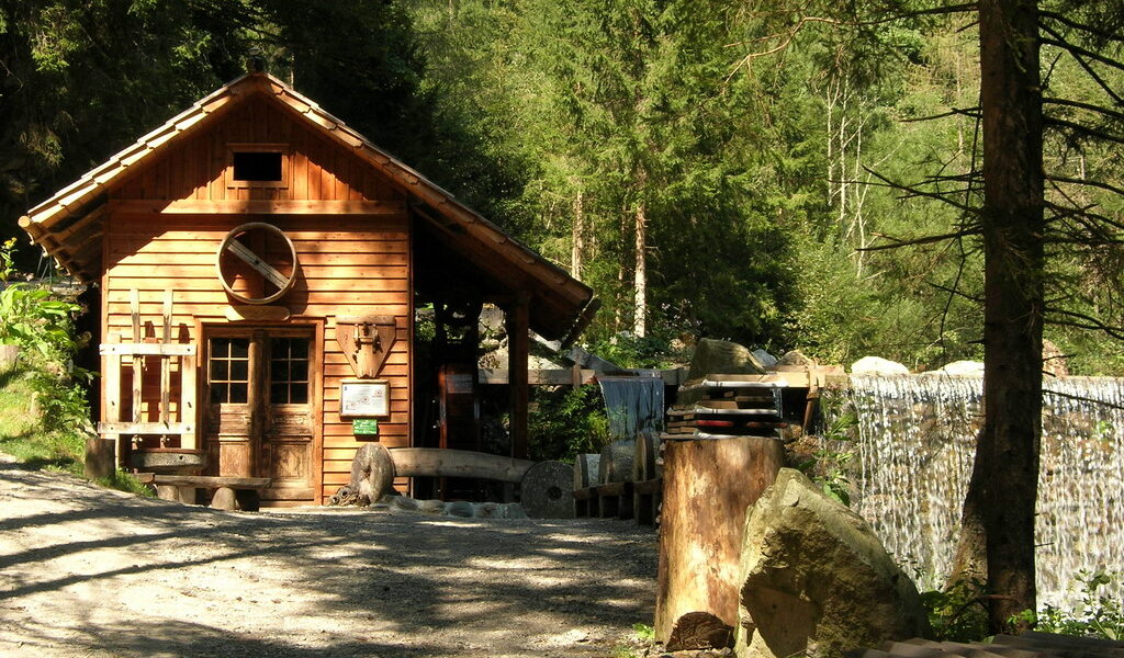 Eine kleine Holzhütte steht neben einem Kiesweg in einem Waldgebiet. Das Sonnenlicht bricht durch die Bäume und rechts ist ein kleiner Wasserfall zu sehen.