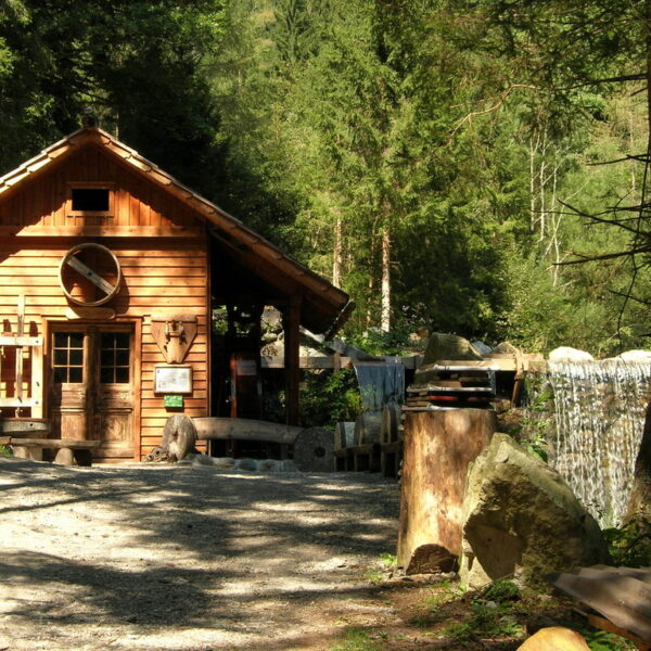 Eine kleine Holzhütte steht neben einem Kiesweg in einem Waldgebiet. Das Sonnenlicht bricht durch die Bäume und rechts ist ein kleiner Wasserfall zu sehen.