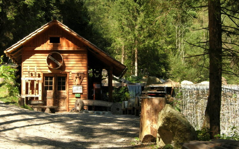 Eine kleine Holzhütte steht neben einem Kiesweg in einem Waldgebiet. Das Sonnenlicht bricht durch die Bäume und rechts ist ein kleiner Wasserfall zu sehen.