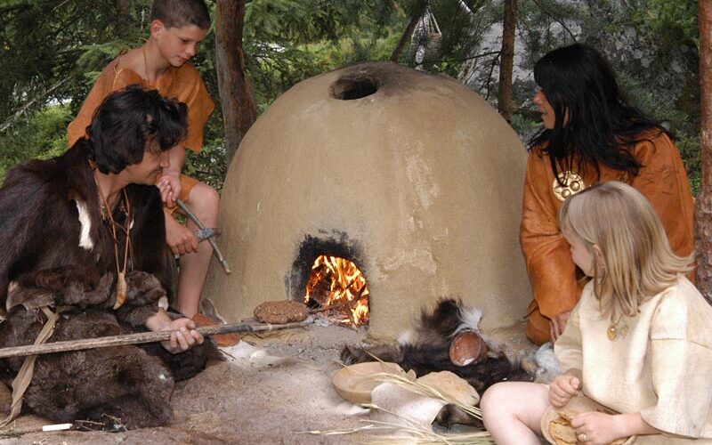 Vier Personen in historischer Kleidung backen Brot an einem Lehmofen im Freien, umgeben von Bäumen und belaubten Ästen.