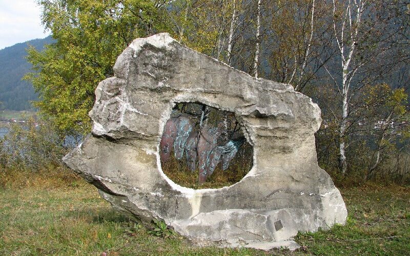 Ein großer, verwitterter Felsen mit einem natürlichen, kreisrunden Loch steht auf einem grasbewachsenen Boden, mit Bäumen und Hügeln im Hintergrund.
