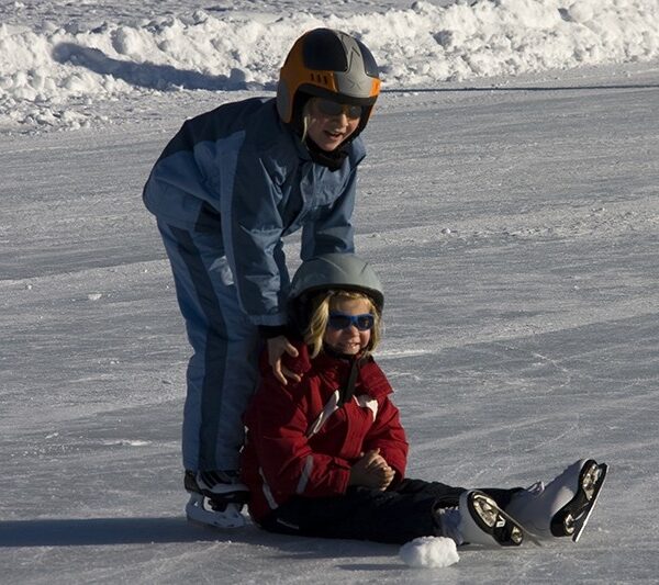 Zwei Kinder, die Helme und Winterkleidung tragen, laufen Schlittschuh; das eine steht und hilft dem anderen, das auf dem Eis sitzt.