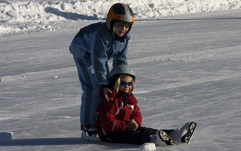 Zwei Kinder, die Helme und Winterkleidung tragen, laufen Schlittschuh; das eine steht und hilft dem anderen, das auf dem Eis sitzt.