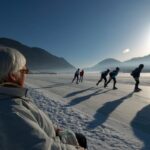 Eine ältere Person sitzt auf einer verschneiten Bank und beobachtet eine Gruppe von Menschen, die auf einem zugefrorenen See, umgeben von Bergen, im Sonnenlicht Schlittschuh laufen.