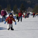 Eine Gruppe von Menschen, die an einem verschneiten Tag im Freien Schlittschuhlaufen, mit einem Kind im Vordergrund, das einen Helm, Handschuhe und Winterkleidung trägt. Im Hintergrund sind Bäume und Berge zu sehen.
