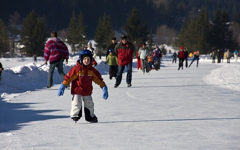 Eine Gruppe von Menschen, die an einem verschneiten Tag im Freien Schlittschuhlaufen, mit einem Kind im Vordergrund, das einen Helm, Handschuhe und Winterkleidung trägt. Im Hintergrund sind Bäume und Berge zu sehen.