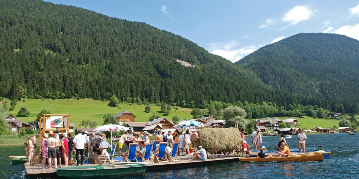 Eine Gruppe von Menschen steht und sitzt auf einem großen Holzfloß mit Liegestühlen und Sonnenschirmen auf einem See, umgeben von grünen Hügeln und Häusern.