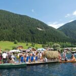 Eine Gruppe von Menschen steht und sitzt auf einem großen Holzfloß mit Liegestühlen und Sonnenschirmen auf einem See, umgeben von grünen Hügeln und Häusern.
