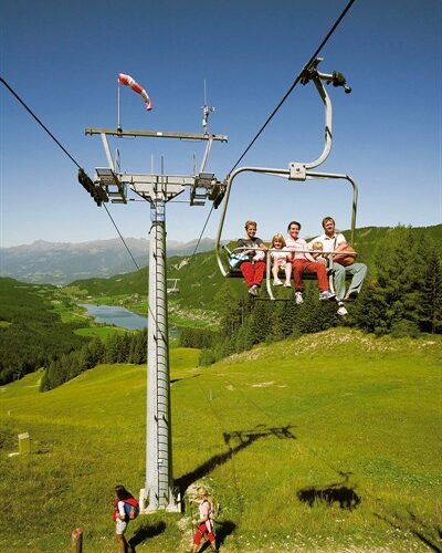 Vier Personen fahren mit einem Sessellift über einen grünen Hügel mit einem See und Bergen im Hintergrund; zwei Wanderer gehen unter ihnen auf dem Gras.