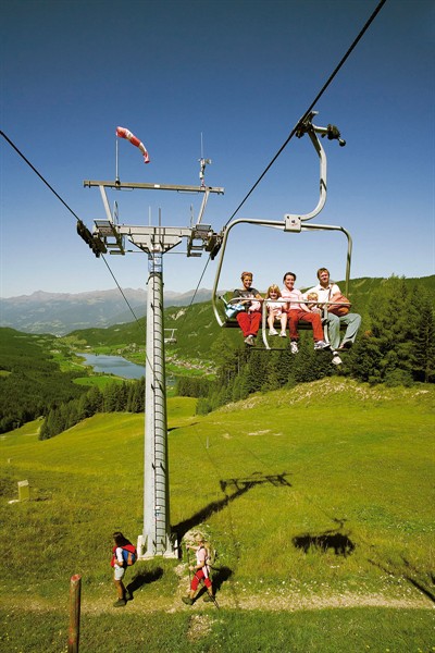 Vier Personen fahren mit einem Sessellift über einen grünen Hügel mit einem See und Bergen im Hintergrund; zwei Wanderer gehen unter ihnen auf dem Gras.