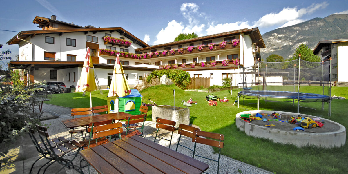 Sitzbereich im Freien mit Holztischen und -bänken neben einem grasbewachsenen Hof mit Spielplatz, Sandkasten, Trampolin und einem Gebäude im Alpenstil im Hintergrund.