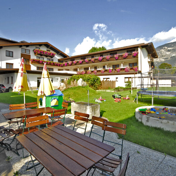 Sitzbereich im Freien mit Holztischen und -bänken neben einem grasbewachsenen Hof mit Spielplatz, Sandkasten, Trampolin und einem Gebäude im Alpenstil im Hintergrund.