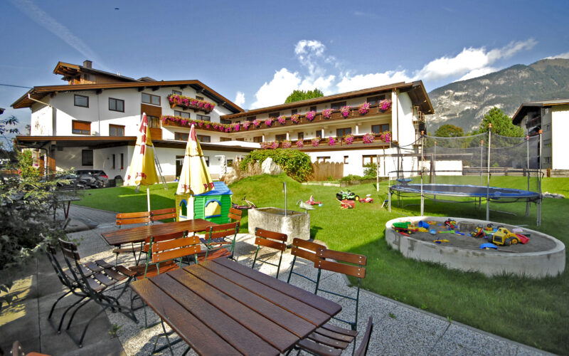Sitzbereich im Freien mit Holztischen und -bänken neben einem grasbewachsenen Hof mit Spielplatz, Sandkasten, Trampolin und einem Gebäude im Alpenstil im Hintergrund.
