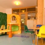 Ein hell erleuchtetes Spielzimmer mit einer Spielstruktur aus Kunststoff, bunten Stühlen um einen Tisch und einem an der Wand befestigten Perlenlabyrinth.