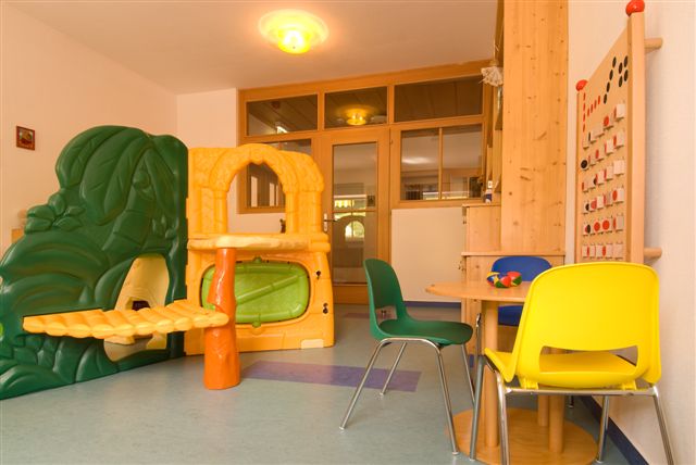 Ein hell erleuchtetes Spielzimmer mit einer Spielstruktur aus Kunststoff, bunten Stühlen um einen Tisch und einem an der Wand befestigten Perlenlabyrinth.