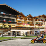 Ein kleines Kind fährt mit einem Tret-Gokart auf einem gepflasterten Weg vor einem großen Hotel im Alpenstil mit gelben Sonnenschirmen und Blumenkästen.