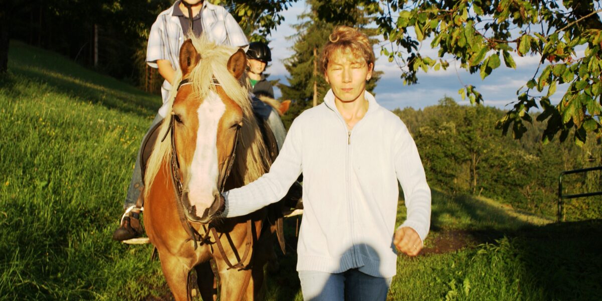 Eine Person führt ein hellbraunes Pferd mit blonder Mähne, während ein Kind mit einem Helm auf dem Rücken des Pferdes auf einer sonnenbeschienenen Wiese reitet.