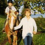 Eine Person führt ein hellbraunes Pferd mit blonder Mähne, während ein Kind mit einem Helm auf dem Rücken des Pferdes auf einer sonnenbeschienenen Wiese reitet.